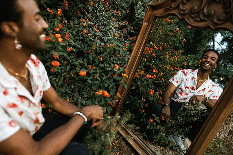 A man kneels next to a bed of flowers, smiling at his reflection in a mirror.