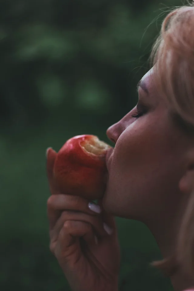 A woman biting into a red apple, surrounded by greenery.