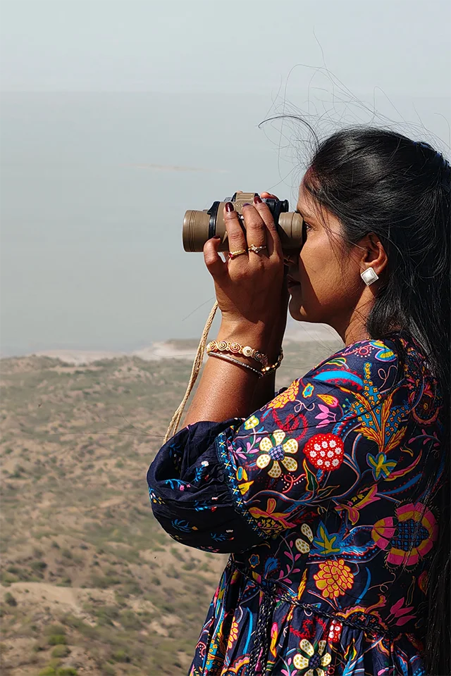 A woman scanning the horizon through binoculars from a hilltop.