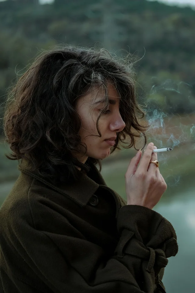 A woman stands by a lake on an overcast day, smoking a cigarette.